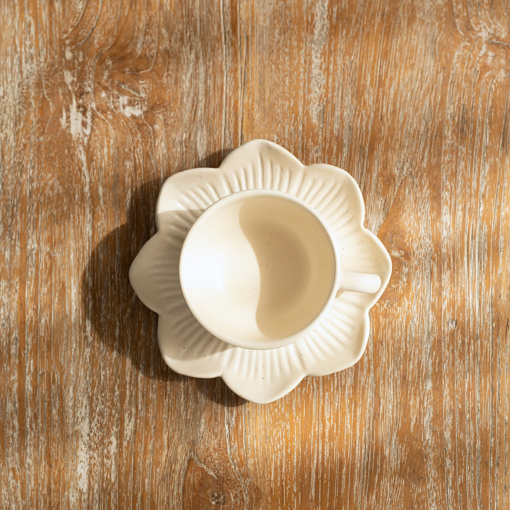 A beige ceramic tea cup placed on a saucer, which is designed to resemble a sunflower, set on a wooden surface.