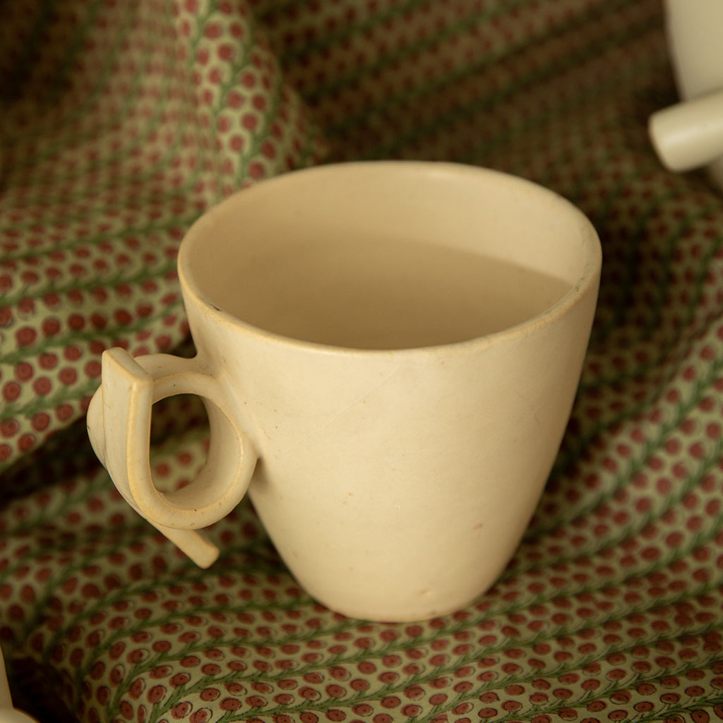 A white ceramic mug with a knotted handle, placed on a patterned fabric.