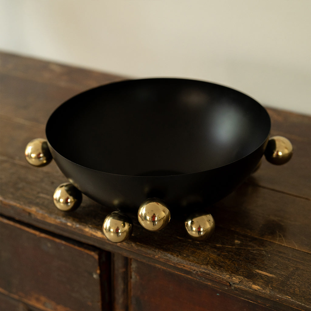 A black decorative stainless steel bowl with gold-colored bubble-shaped handles on a wooden surface.