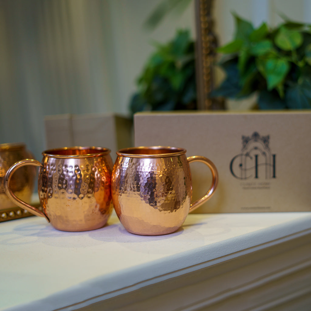 Two copper mule mugs with a shiny finish, placed on a surface with a cardboard box in the background of Curate Home brand.