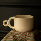 A white ceramic mug with a double looped handle, placed on a patterned fabric, against a dark background.