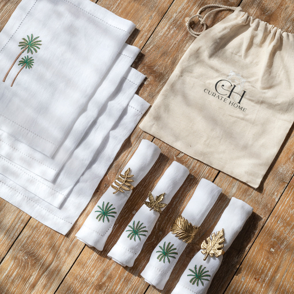 A set of white linen table mats, napkins, and napkin rings displayed on a wooden surface, with a few pieces featuring green palm tree embroidery. A beige cotton drawstring bag with the Curate Home Brand is also visible.