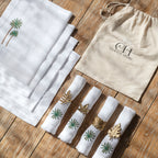 A set of white linen table mats, napkins, and napkin rings displayed on a wooden surface, with a few pieces featuring green palm tree embroidery. A beige cotton drawstring bag with the Curate Home Brand is also visible.