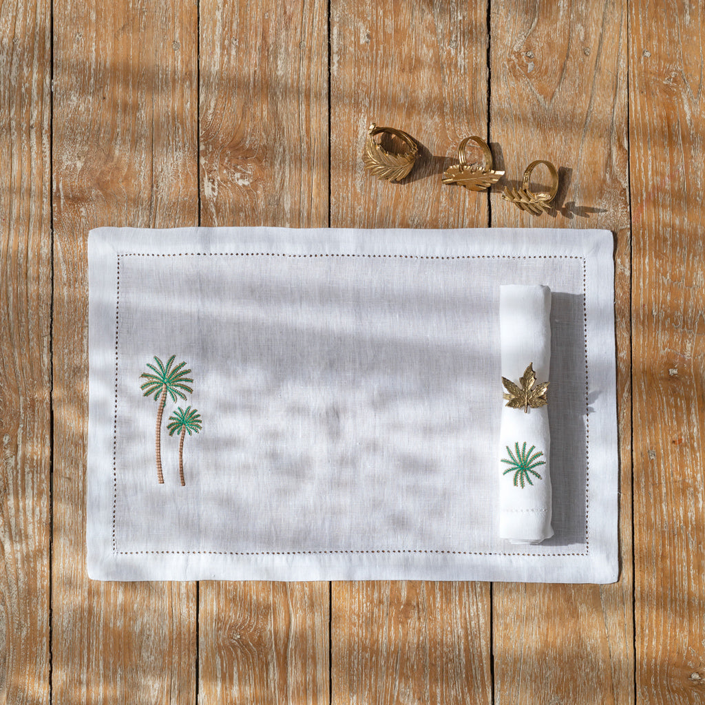 A white linen placemat and napkin with green palm tree embroidery and beige hemstitching, displayed on a wooden table with a pair of gold napkin rings beside it.