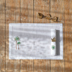 A white linen placemat and napkin with green palm tree embroidery and beige hemstitching, displayed on a wooden table with a pair of gold napkin rings beside it.
