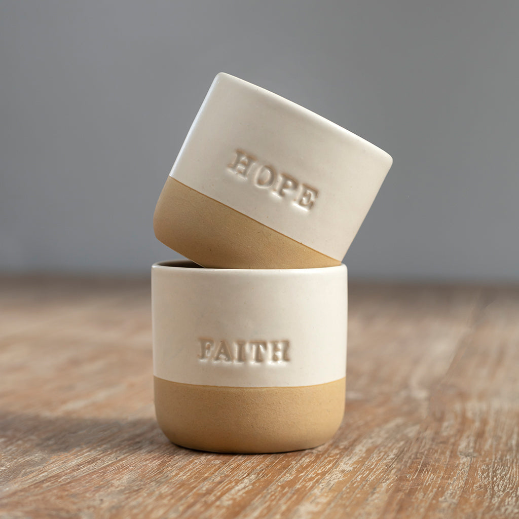 Two beige ceramic cups with the words 'HOPE' and 'FAITH' engraved on them, placed on a wooden surface.