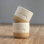 Two beige ceramic cups with the words 'HOPE' and 'FAITH' engraved on them, placed on a wooden surface.