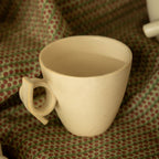 A white ceramic mug with a knotted handle, placed on a patterned fabric.