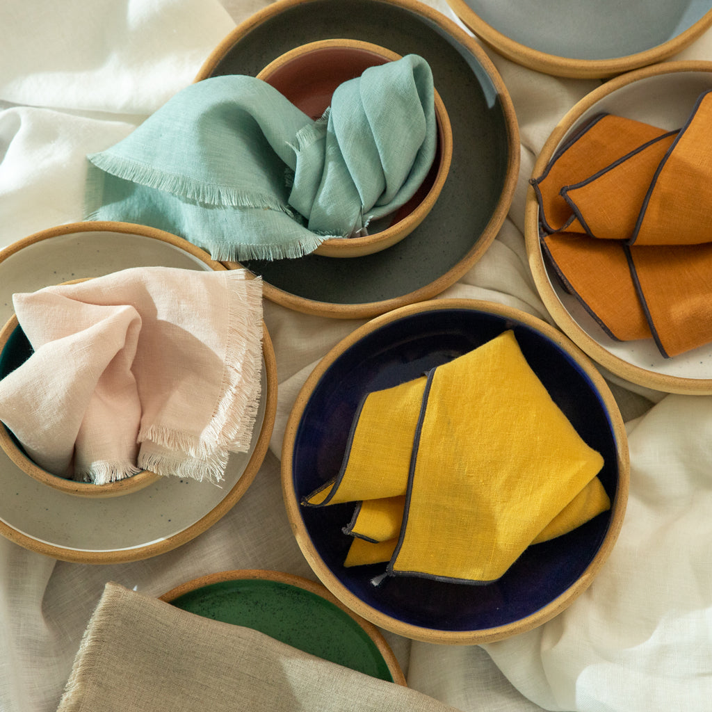 A Woven of 100% linen, these napkins have a raw fringe in various colors, including light pink and light blue.