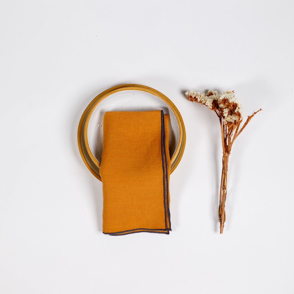 A folded orange linen napkin with a decorative hemstitch, displayed next to a withered flower on a white background. Linen napkin top stitch clay color.