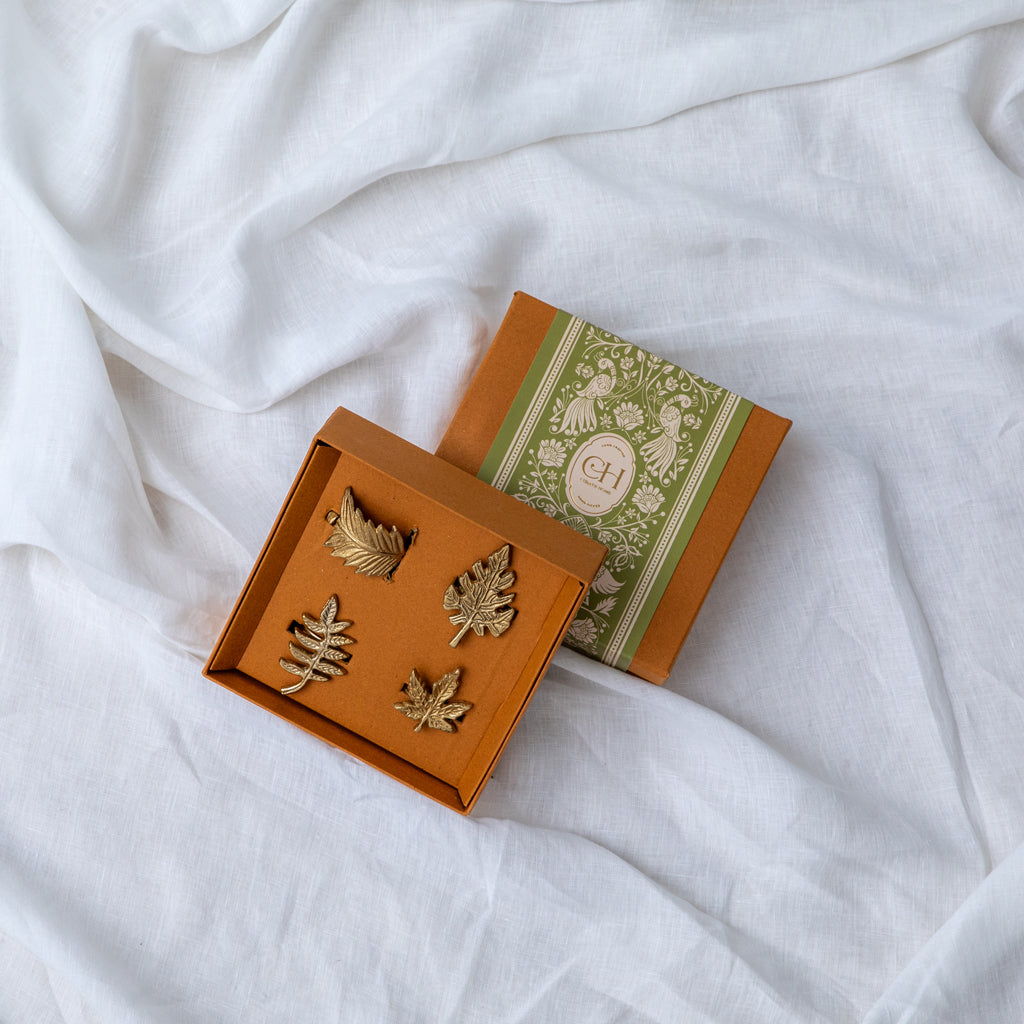 A set of four gold-colored metal napkin rings with a leaf design, displayed in an open box with a decorative pattern.