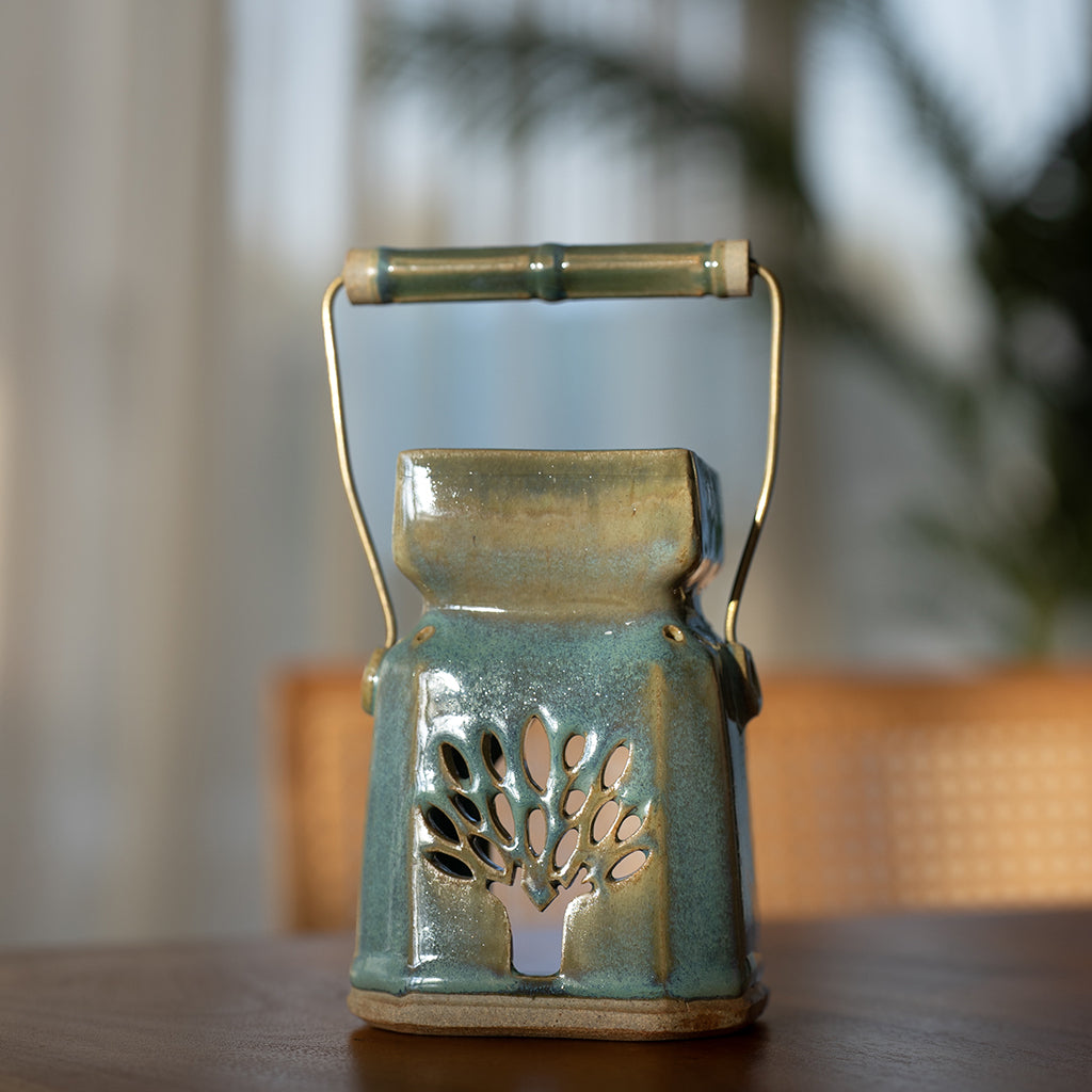 A handmade ceramic palm-shaped diffuser with a brass wire handle, containing a bowl for water and essential oils, with shades of green and brown, placed on a wooden surface.