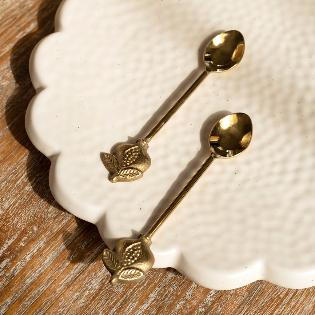 A set of two gold-colored stainless steel pomegranate dessert spoons with leaf patterns on the handles, placed on a white plate.
