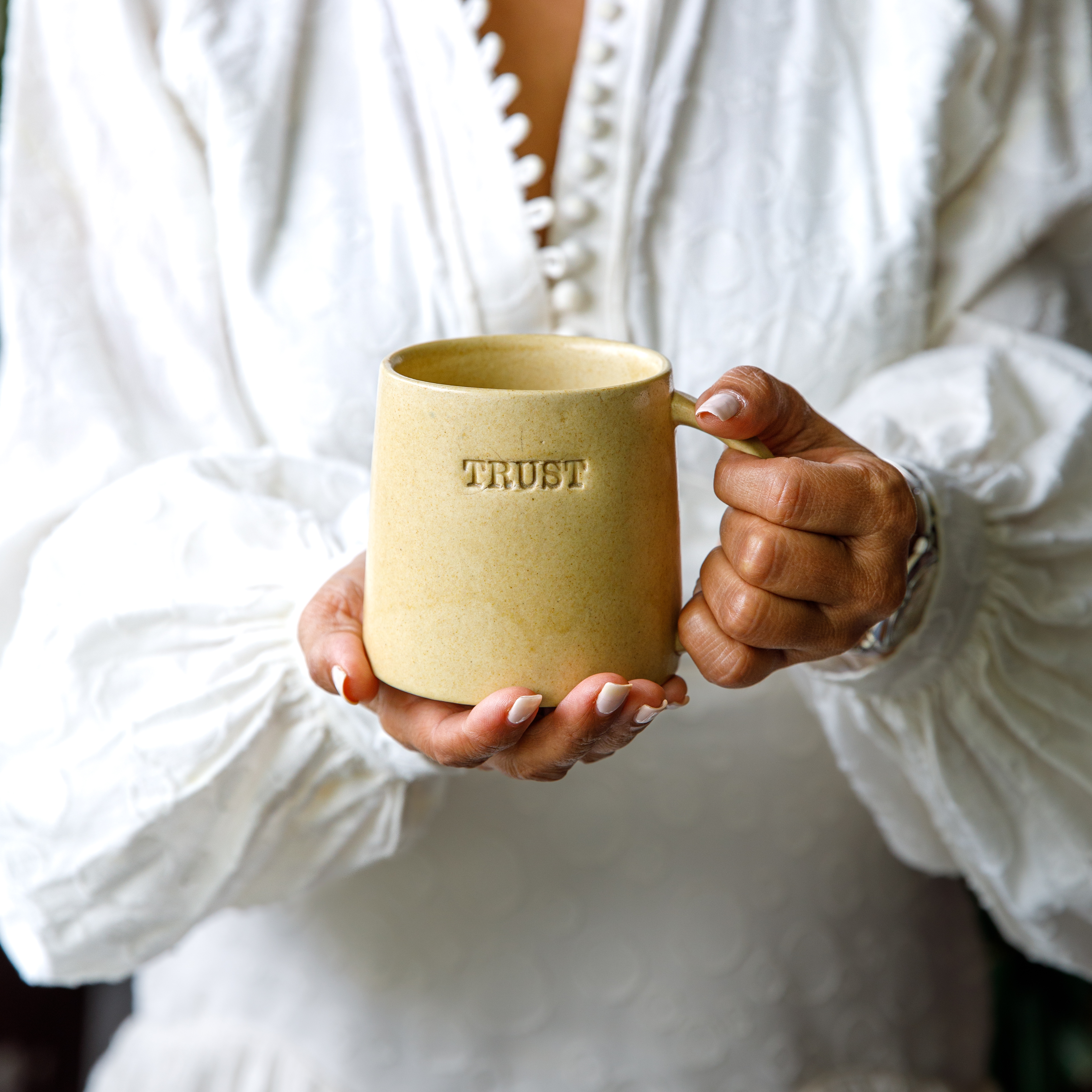 A person holding a beige ceramic mug with the word 'TRUST' engraved on it.