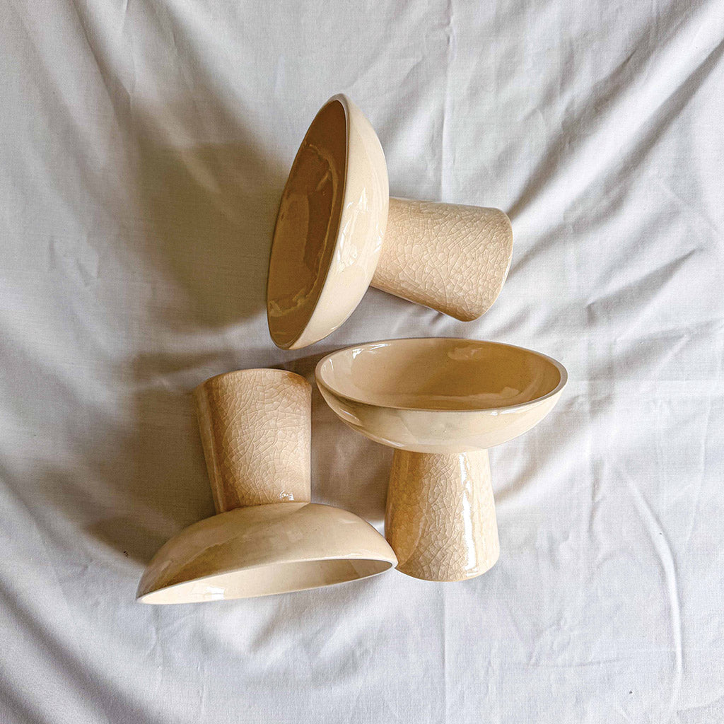 Four cream-colored ceramic tableware pieces with a glossy, crackled glaze, arranged on a wrinkled white fabric background.