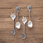 A set of four silver-colored dessert spoons with star and moon designs, placed on a wooden surface. Length 14.6cm.