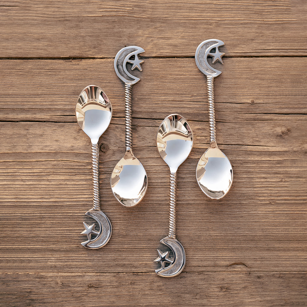 A set of four silver-colored dessert spoons with star and moon designs, placed on a wooden surface. Length 14.6cm.