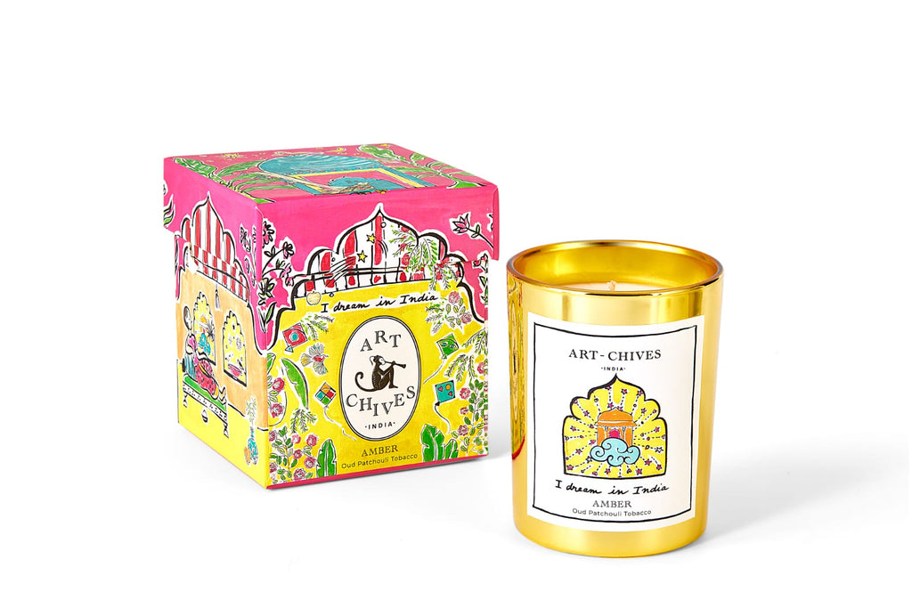 A fragranced made with 100% soy candle with luxe packaging, showcased next to its vibrant pink box adorned with decorative elements and the Art Chives brand label. The Amber Candle